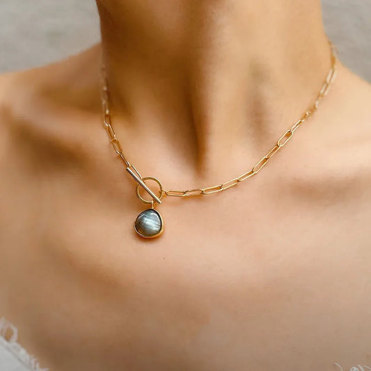 Detailaufnahme der 18K vergoldeten Messing-Gliederkette mit Kristall-Anhänger und Toggle-Verschluss, getragen zu einem weißen, ausgeschnittenen Oberteil. Die Kette liegt nah am Hals.