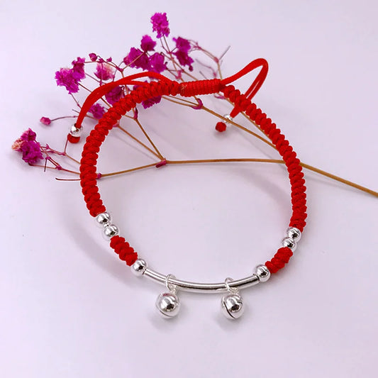 Handgeflochtenes rotes Glücksarmband mit silbernen Glöckchen dekorativ neben violetten Trockenblumen arrangiert.