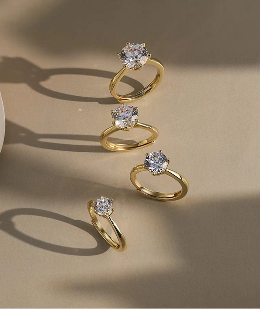 Vier goldfarbene, verstellbare Ringe aus vergoldetem 925er Silber, jeweils mit einem einzelnen, runden Zirkonia-Stein besetzt, auf hellem Untergrund arrangiert.