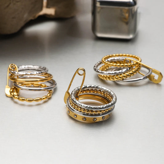 Mehrere Ringe, bestehend aus gestapelten goldenen und silbernen Ringen, liegen auf einer hellen Oberfläche. Die Ringe haben einen dekorativen Sicherheitsnadel-Verschluss.