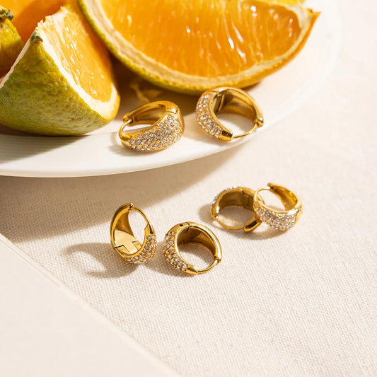 Zwei kegelförmige Creolen und zwei runde Creolen aus 18K vergoldetem Edelstahl mit Zirkonia-Steinen auf einem Teller mit Orangenscheiben.