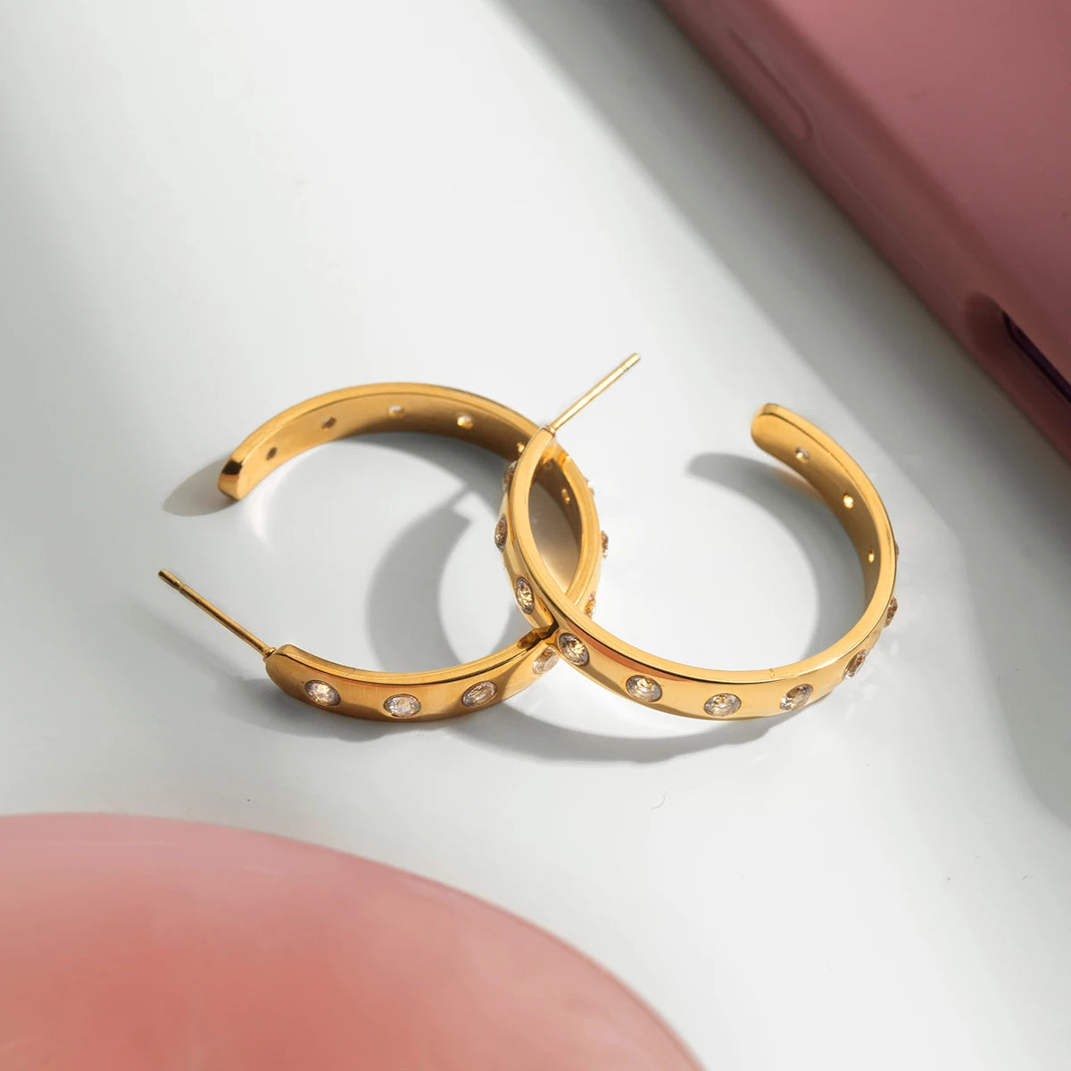Zwei goldene Chunky-Ohrringe aus Edelstahl mit glitzernden Steinen, flach liegend im hellen Sonnenlicht mit harten Schatten.