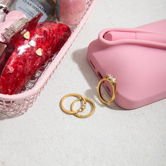 Set aus drei goldenen Ringen mit Zirkoniasteinen, neben einer pinken Schlafmaske und in einer weichen, pinken Schale, was eine gemütliche Abendroutine suggeriert.