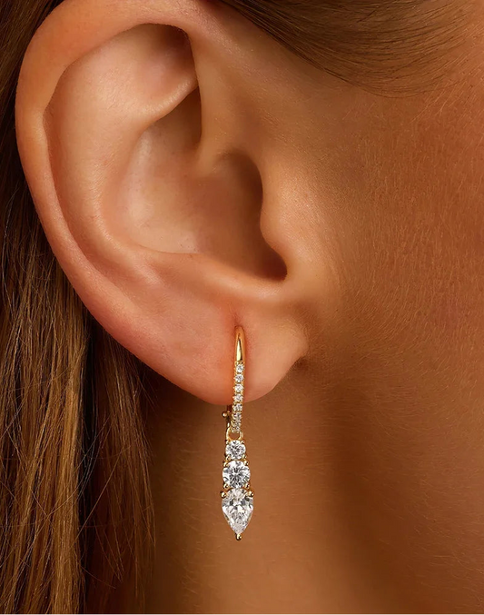 Nahaufnahme eines weiblichen Ohrs, das einen eleganten, hängenden Ohrring aus 14K vergoldetem 925er Silber trägt. Der Ohrring ist mit drei tropfenförmigen, gestuften Zirkonia-Steinen und einem kleinen Zirkonia-besetzten Kreolenbügel verziert.