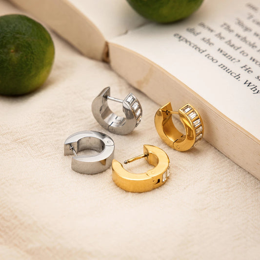 Elegantes Stillleben-Foto von vier Huggie-Klappcreolen aus Edelstahl, die paarweise (Silber und 18K goldvergoldet) auf einem hellen Podest neben einem offenen Buch und zwei Limetten liegen, alle mit Baguette-Zirkonia-Besatz.