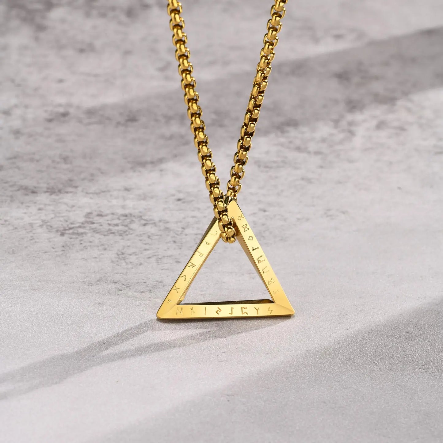 Nahaufnahme der goldfarbenen Edelstahlkette mit dreieckigem Anhänger, wobei die Gravur/Prägung auf dem unteren Rand des Dreiecks sichtbar ist, auf hellem Hintergrund.