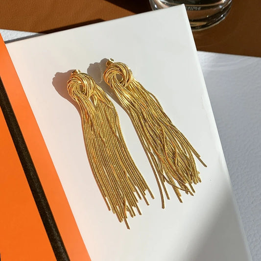 Lange vergoldete Quasten-Ohrringe aus Titan mit gedrehtem Knoten-Detail, aufgeschlagen auf einem Notizbuch.