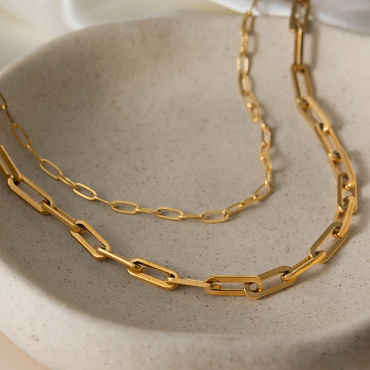 Nahaufnahme des geschichteten Gliederkettensets aus 18K goldvergoldetem Edelstahl, kunstvoll arrangiert in einer hellen, mattierten Steingutschale, um die Textur und den Glanz der Paperclip-Kettenglieder zu zeigen.