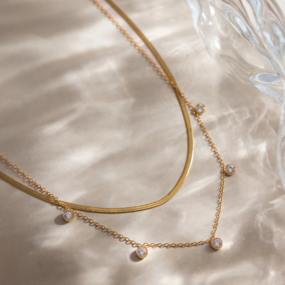 Eine Nahaufnahme des geschichteten Halskettensets aus goldfarbenem Edelstahl, präsentiert auf einem hellen, gewellten Seidenhintergrund; das Set besteht aus einer feinen Gliederkette mit mehreren klaren, brillant-geschliffenen Zirkoniasteinen im Lünettenrahmen und einer breiteren Gliederkette mit 18K Goldvergoldung.