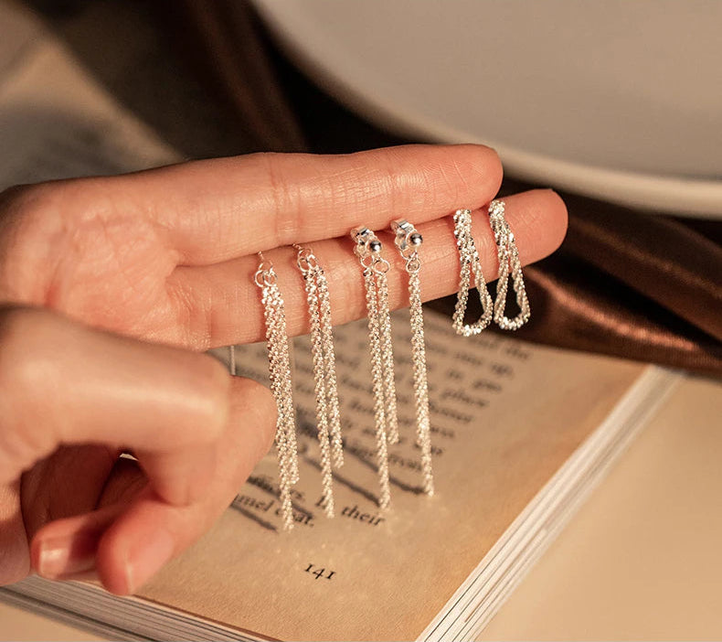 Detailaufnahme von drei Paaren silberfarbener Ohrringe aus 925er Silber – zwei Paare lange Faden-Ohrringe und ein Paar kurze, U-förmige Ohrringe – die auf einem geöffneten Buch präsentiert und von einer Hand gehalten werden.