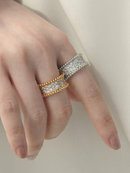 Ein offener Ring aus 18K vergoldetem Messing steht auf einer hellen Fläche; das Sonnenlicht betont die feine Körnung des Goldes und die präzise gefassten Zirkonia-Steine in der Mitte.