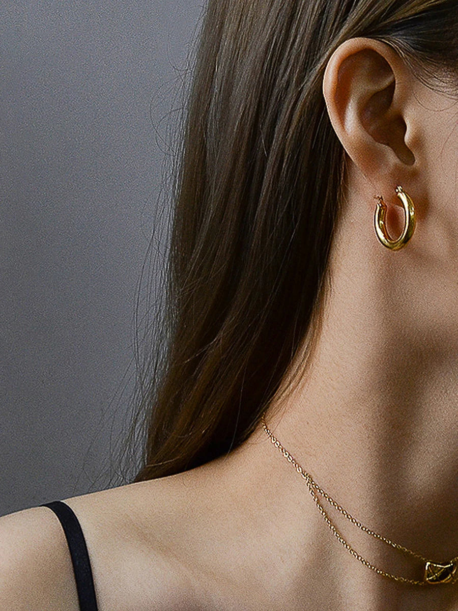 Nahaufnahme von goldenen 18K vergoldeten Creolen und einer zarten Halskette am Ohr einer Frau.