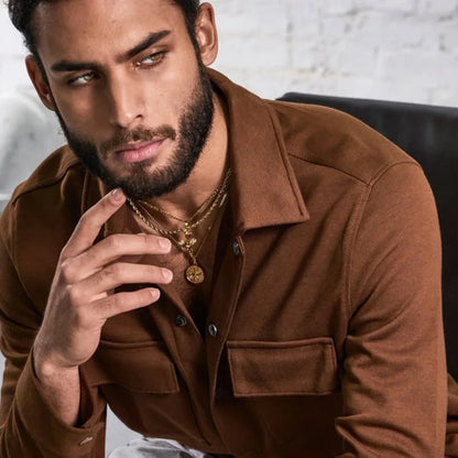 Mann mit Bart trägt zwei goldfarbene Ketten, wobei die untere ein Kompass-Medaillon zeigt, über einem braunen Hemd.