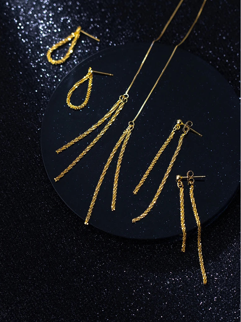 Arrangement von goldfarbenem Schmuck auf einem glitzernden schwarzen Hintergrund, bestehend aus einem Paar einfacher Creolen, einer langen Kette und zwei Paaren langer Faden-Ohrringe (Material: 925er Silber).