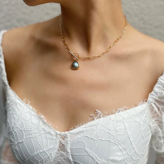 Eine Frau trägt die kurze, 18K vergoldete Messing-Gliederkette mit Kristall-Anhänger über einem weißen, rüschenbesetzten Oberteil. Halbkörperansicht.