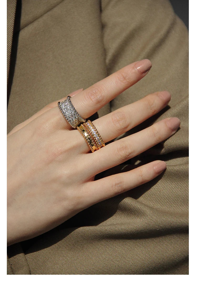 Detailansicht einer Hand, an der zwei strukturierte Ringe übereinander getragen werden; das Design kombiniert den warmen Glanz der 18K Vergoldung mit dem brillanten Funkeln zahlreicher kleiner Zirkonia.