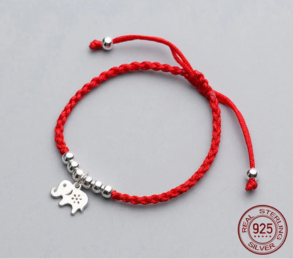 Nahaufnahme eines geflochtenen roten Textilarmbands mit kleinen Silberperlen und verziertem Elefanten-Anhänger aus 925er Sterling Silber auf grauem Hintergrund.