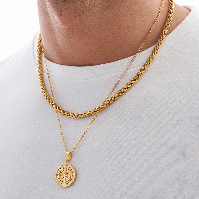 Mann trägt zwei goldfarbene Edelstahlketten, eine geflochtene und eine mit Kompass-Medaillon, über einem weißen T-Shirt.