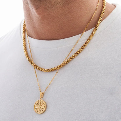Mann trägt zwei goldfarbene Edelstahlketten, eine geflochtene und eine mit Kompass-Medaillon, über einem weißen T-Shirt.