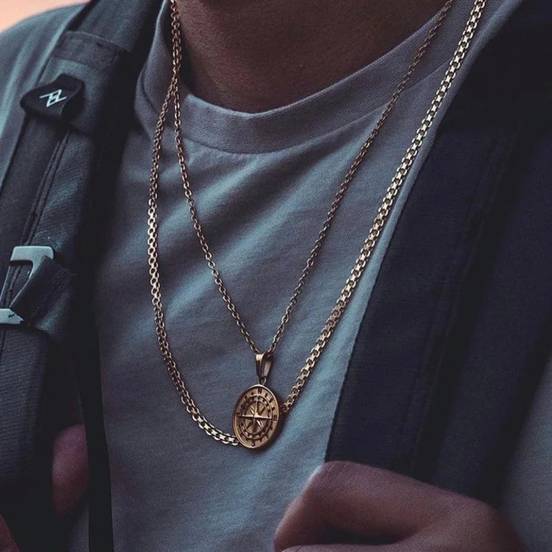 Nahaufnahme eines Mannes, der zwei goldfarbene Edelstahlketten übereinander trägt: eine engmaschige und eine mit einem ovalen Kompass-Medaillon. Die Ketten werden über einem hellgrauen T-Shirt gezeigt.