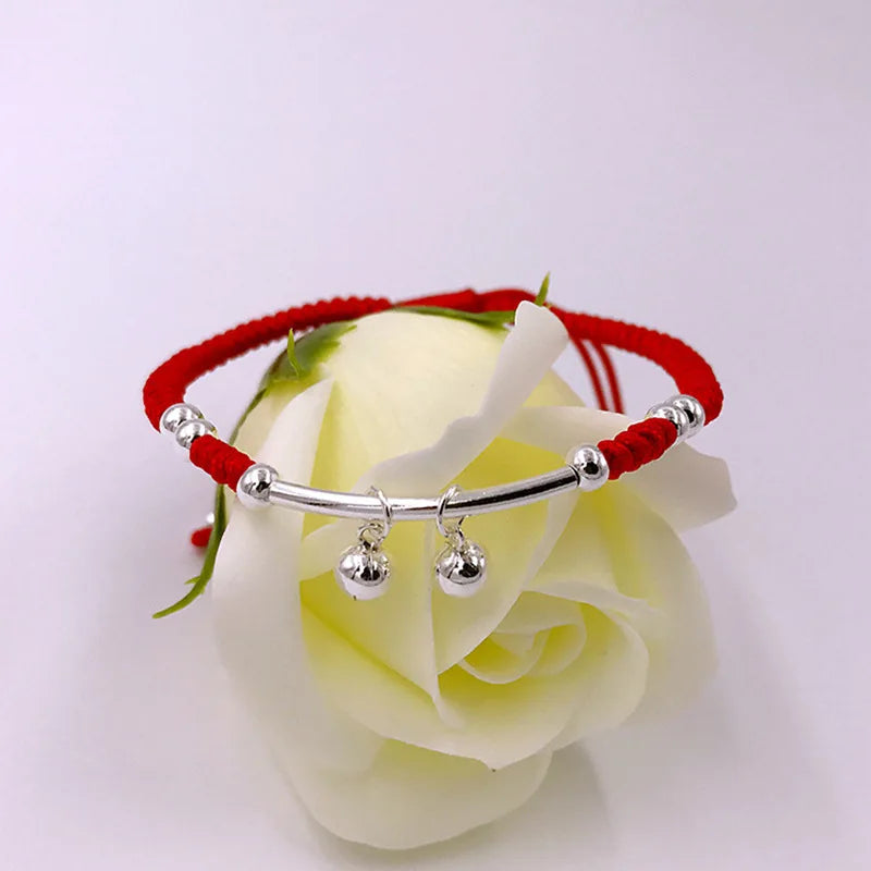 Produktansicht des roten Kordelarmbands mit zwei Glöckchen-Anhängern elegant auf einer weißen Rose platziert.