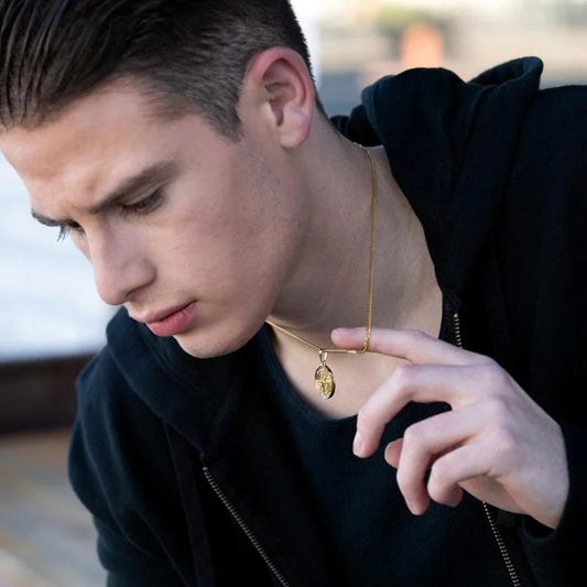 Junger Mann trägt eine zarte goldfarbene Kette mit kleinem Kompass-Medaillon, bekleidet mit einem dunklen Kapuzenpullover.