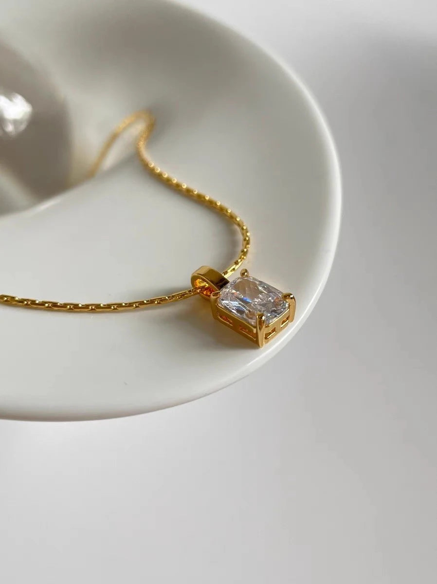 Nahaufnahme einer Frau, die die goldene Kette mit dem rechteckigen Zirkonia-Anhänger trägt; dazu kombiniert sie ein schwarzes Top, eine weiße Bluse und einen passenden Goldring.