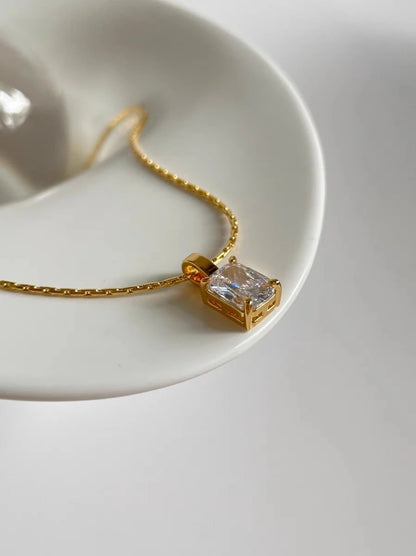 Nahaufnahme einer Frau, die die goldene Kette mit dem rechteckigen Zirkonia-Anhänger trägt; dazu kombiniert sie ein schwarzes Top, eine weiße Bluse und einen passenden Goldring.