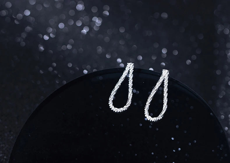 Nahaufnahme eines Paares silberfarbener, U-förmiger Ohrringe aus 925er Silber, die mit kleinen, glitzernden Steinen besetzt sind, präsentiert auf einem glänzenden, schwarzen Podest.