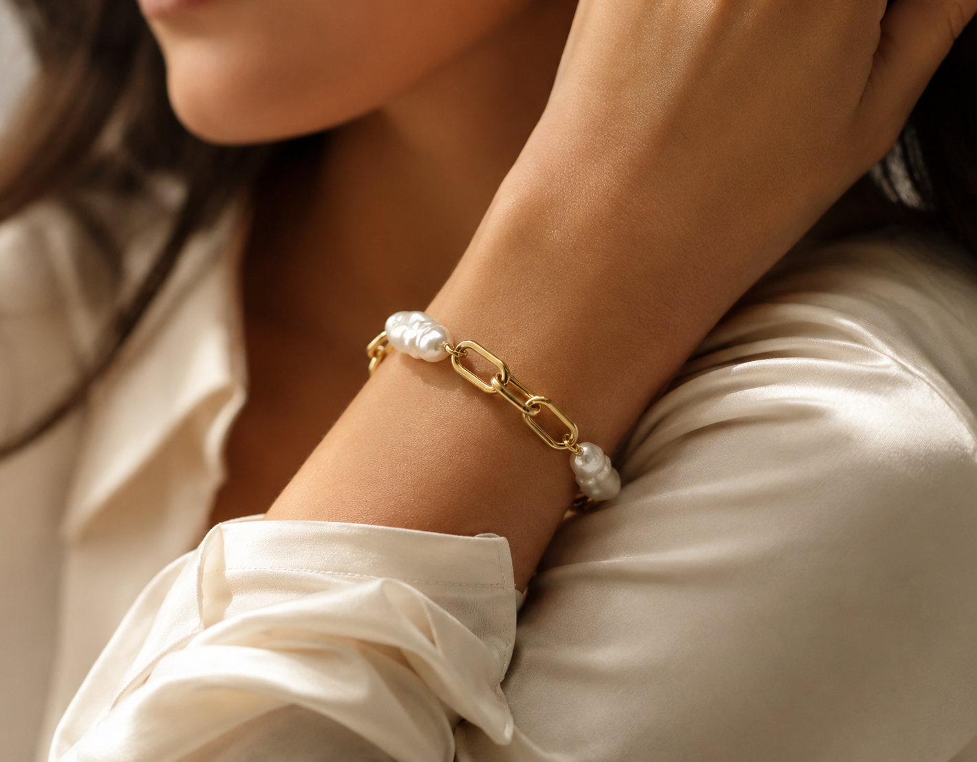 Elegantes Schmuck-Detail am Handgelenk: Eine Frau trägt das goldene Gliederarmband mit echten Barockperlen zu einer cremefarbenen Seidenbluse.