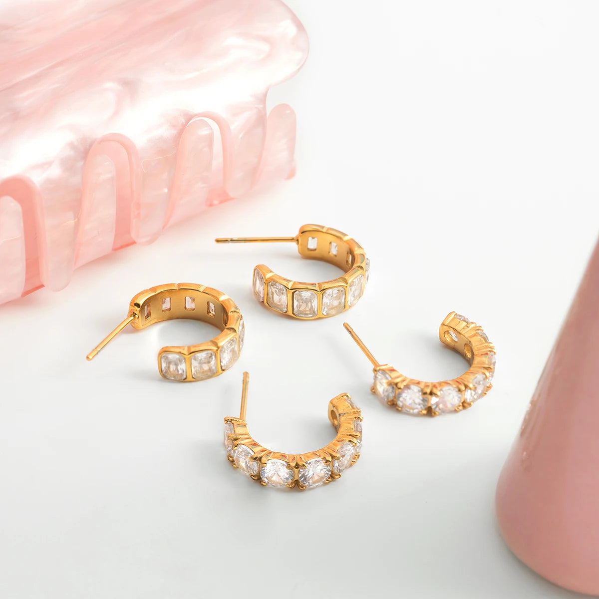 Stilvolle Produktfotografie mehrerer goldener Edelstahl-Creolen mit Zirkonia-Besatz auf heller Oberfläche neben rosafarbenen Accessoires.