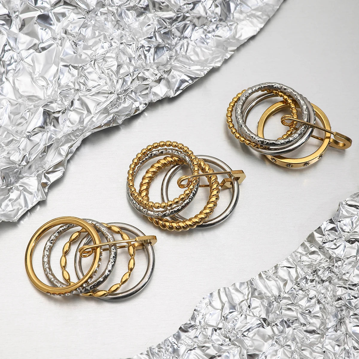 Mehrere Ringe mit dem gestapelten goldenen und silbernen Design, das einen dekorativen Sicherheitsnadel-Verschluss hat, liegen auf zerknitterter silberner Folie.