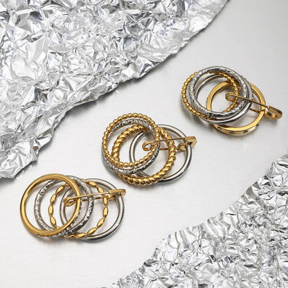 Mehrere Ringe mit dem gestapelten goldenen und silbernen Design, das einen dekorativen Sicherheitsnadel-Verschluss hat, liegen auf zerknitterter silberner Folie.