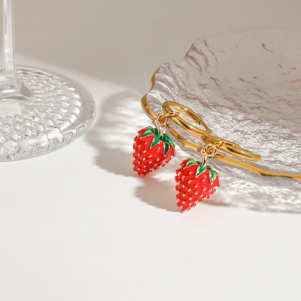 Sommerlicher Schmuck: 18K vergoldete Creolen mit plastisch geformten, roten Erdbeeren, präsentiert auf einer strukturierten Glasunterlage.