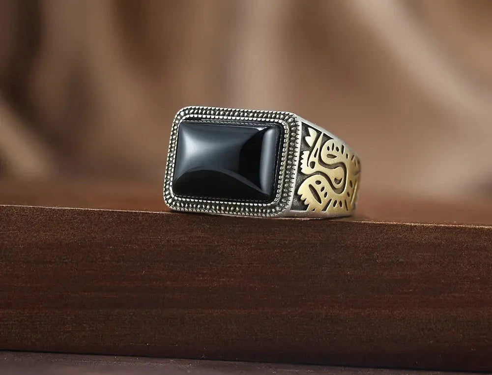 Massiver Herrenring aus oxidiertem 925er Silber mit rechteckigem schwarzen Achat-Stein und goldenen Gravur-Details, platziert auf dunklem Holz.