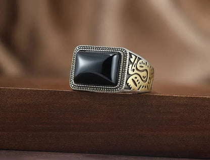 Massiver Herrenring aus oxidiertem 925er Silber mit rechteckigem schwarzen Achat-Stein und goldenen Gravur-Details, platziert auf dunklem Holz.