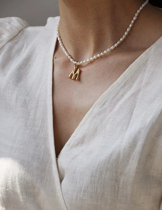 Elegante Perlenkette mit vergoldetem Buchstabe-M-Anhänger am Dekolleté zu einem weißen V-Neck-Oberteil.