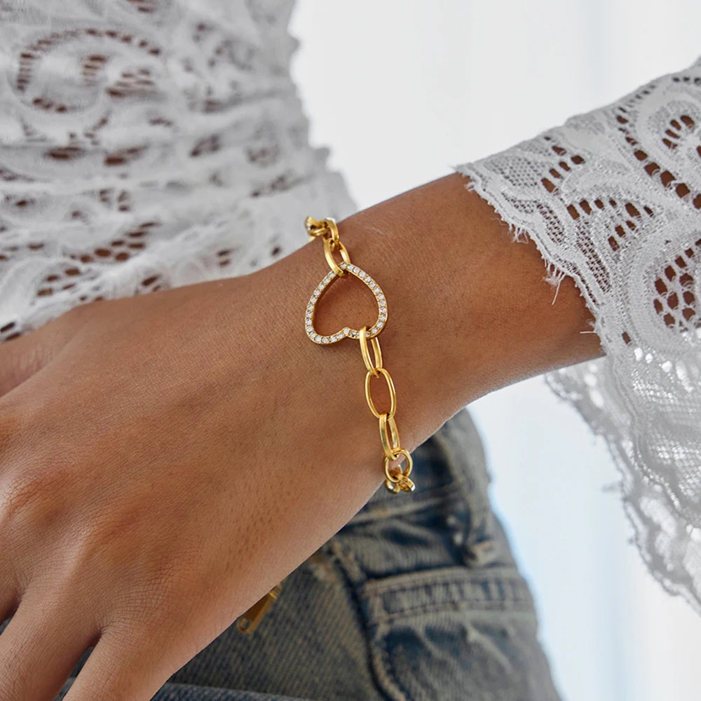 Nahaufnahme am Handgelenk: Gliederarmband aus 18K vergoldetem Edelstahl mit offenem, funkelndem Zirkonia-Herz-Glied zu einer Spitzenbluse