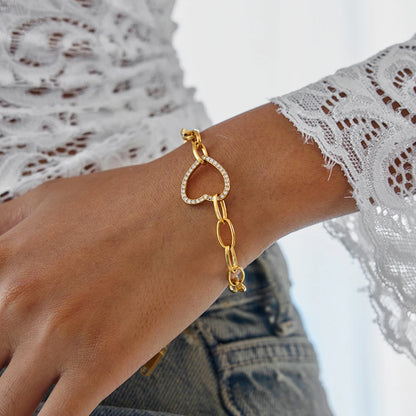 Nahaufnahme am Handgelenk: Gliederarmband aus 18K vergoldetem Edelstahl mit offenem, funkelndem Zirkonia-Herz-Glied zu einer Spitzenbluse