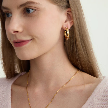 Porträt einer lächelnden Frau, die die 18K vergoldeten Chunky-Creolen mit Zirkonien zu einer feinen Goldkette und einem hellrosa Oberteil kombiniert.