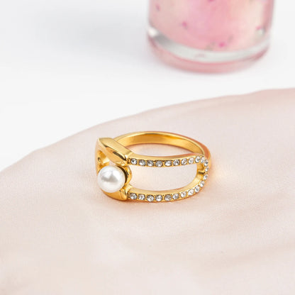 goldfarbener Edelstahlring mit 18K PVD-Vergoldung, Strasssteinen und einer Perle, auf einer weißen Unterlage in der Nähe von Blütenblättern.