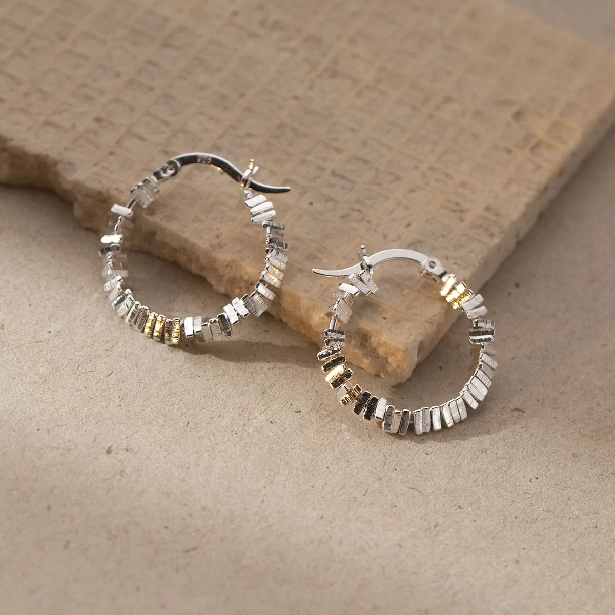  Zwei strukturierte Kreolen-Ohrringe (925er Silber) liegen auf einer hellbraunen, strukturierten Oberfläche (vermutlich Stein oder Karton).