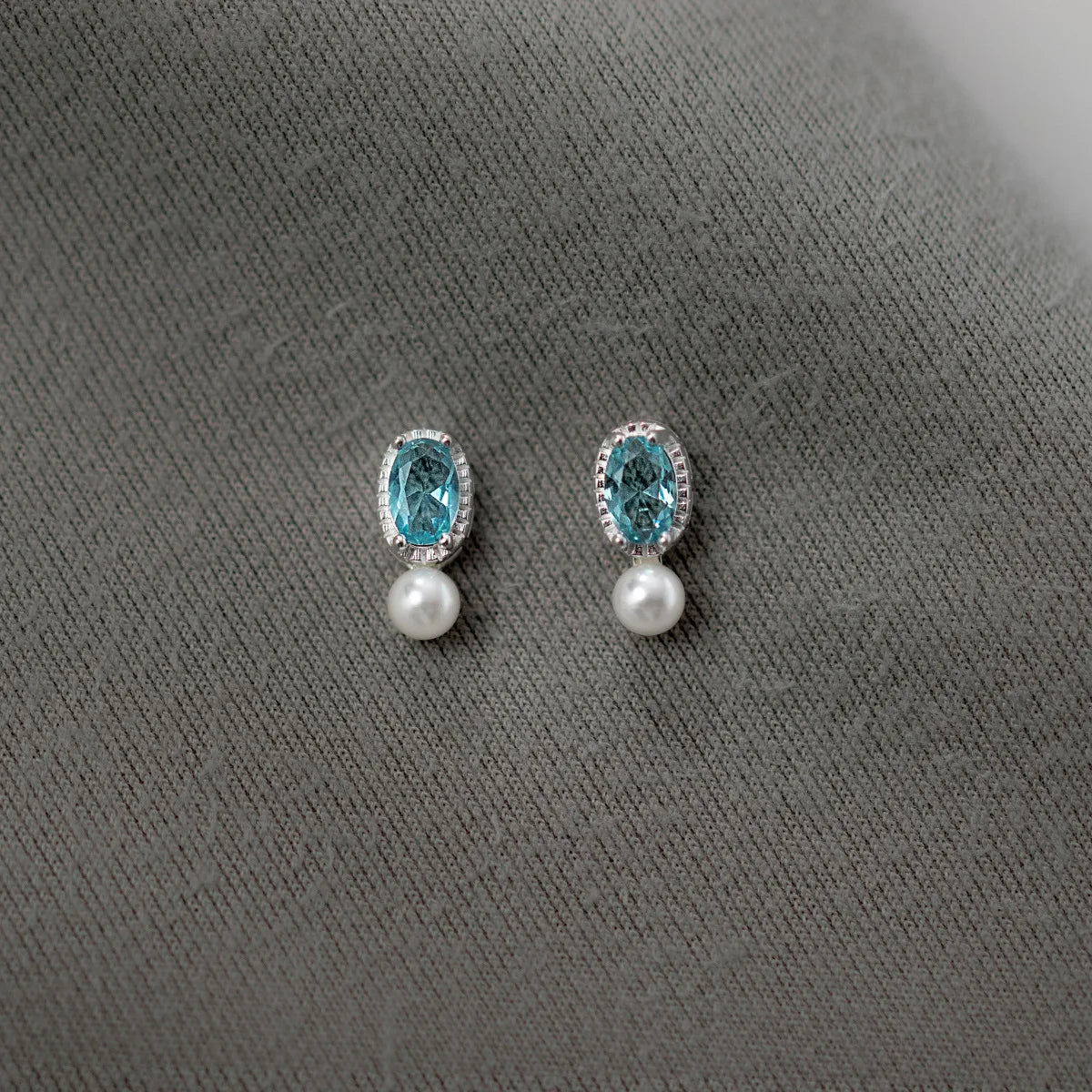 Ein Paar Ohrstecker aus reinem 925er Sterling Silber mit ovalem, aquablauen Zirkoniastein und kleiner weißer Perle, präsentiert auf grauem Stoffhintergrund.