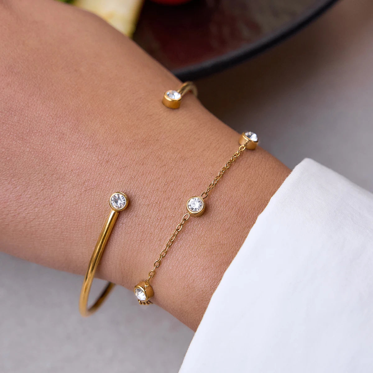 Nahaufnahme eines 2-teiligen Armband-Sets am Handgelenk: Ein offener, 18K vergoldeter Edelstahl-Armreif mit Zirkoniern an den Enden, kombiniert mit einer feinen Goldkette mit runden Kristalleinfassungen.