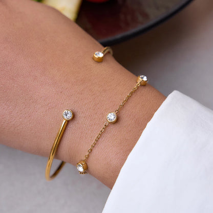 Nahaufnahme eines 2-teiligen Armband-Sets am Handgelenk: Ein offener, 18K vergoldeter Edelstahl-Armreif mit Zirkoniern an den Enden, kombiniert mit einer feinen Goldkette mit runden Kristalleinfassungen.