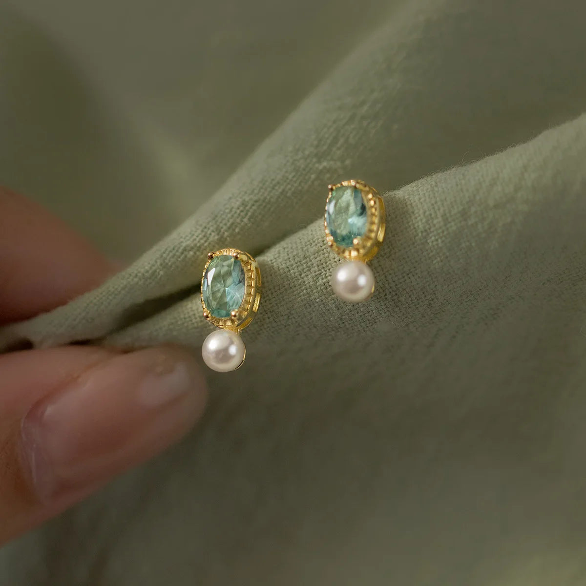 Ein Paar elegante Ohrringe aus 18K vergoldetem 925er Silber mit hellblauem Schmuckstein und kleiner Perle, flach arrangiert auf beigefarbenem Hintergrund.