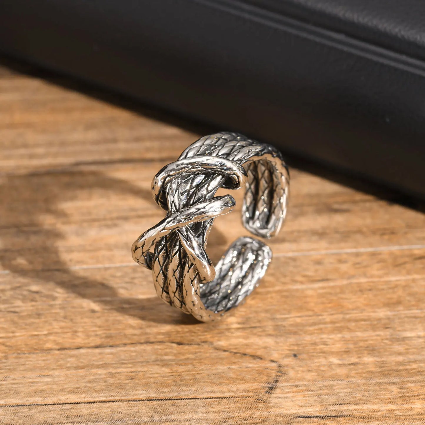 Breiter, offener Edelstahl-Ring im gedrehten Seil- oder Knoten-Design (Tauwerk-Muster), liegend auf einem Holzuntergrund, um das rustikale, oxidierte Silber-Finish hervorzuheben.
