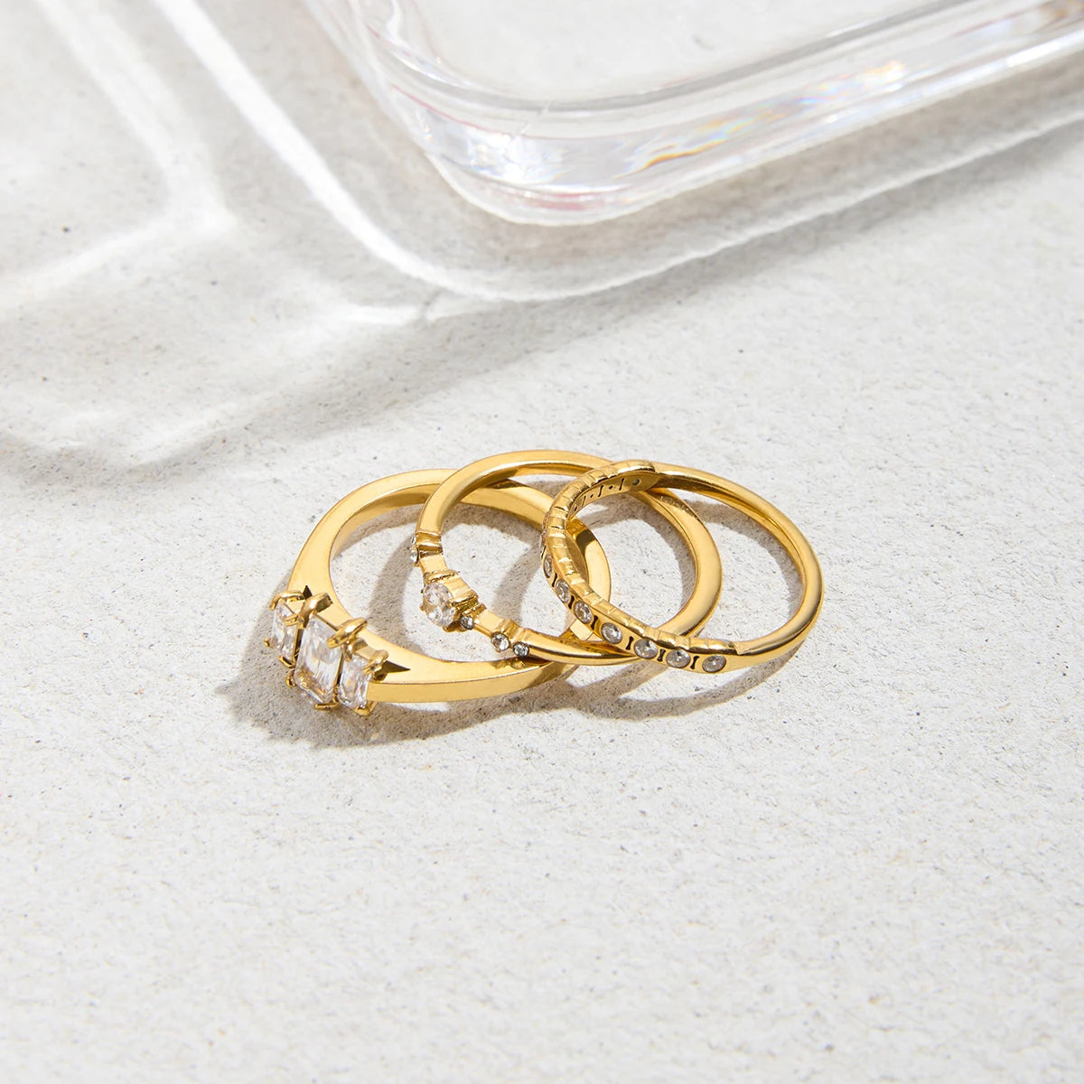 Drei goldene Ringe, kunstvoll gestapelt auf einer reflektierenden Oberfläche. Das Set besteht aus einem Solitärring, einem Memoirering und einem Ring mit drei Zirkonia-Steinen.