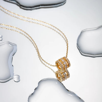 Die vergoldete Edelstahlkette mit Zirkonia-Anhänger liegt auf einer reflektierenden Oberfläche mit Wassertropfen, was das edle Design und die Lichtreflexionen betont.