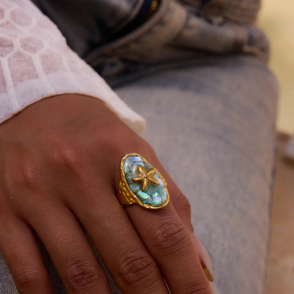 Hand einer Person mit einem ovalen Ring mit türkisfarbenem Stein und Seestern-Symbol aus 18K vergoldetem Edelstahl, mit Meer im Hintergrund.